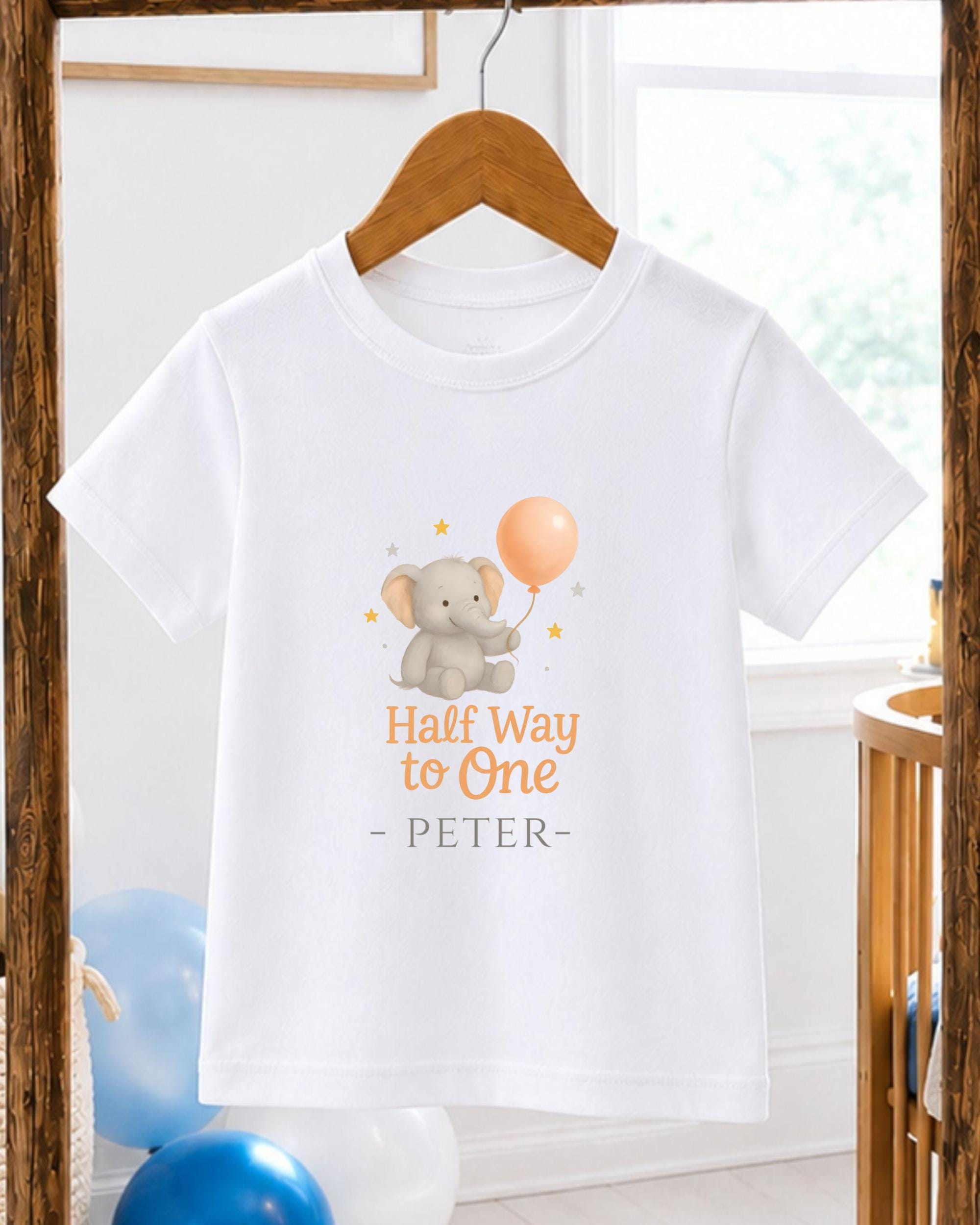 A white t-shirt with a graphic design featuring a gray elephant holding a balloon, along with the text "Half Way to One - PETER".
