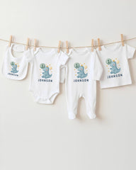 A collection of white baby clothes, including onesies and bibs, with dinosaur designs and the name "Johnson" printed on them.