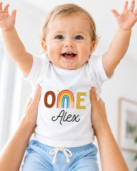 A smiling baby wearing a white t-shirt with the name "Olive" and the name "Alex" printed on it, with their arms raised in the air.
