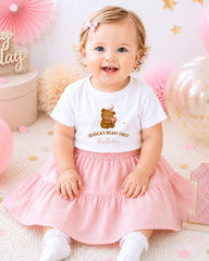 A smiling baby girl wearing a white t-shirt with a teddy bear graphic and the text " Jessica's first birthday" sits on a pink and white background with balloons and confetti.