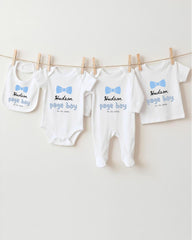 A row of four white baby clothes with blue bows hanging on a clothesline.