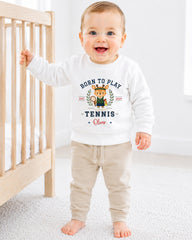 A smiling baby boy wearing a white sweatshirt with a tennis-themed design and text.