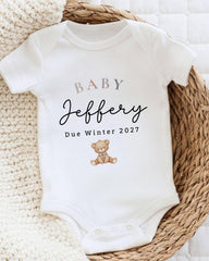 A white baby onesie with the text "BABY Jeffery Due Winter 2027" printed on it, along with a teddy bear image.