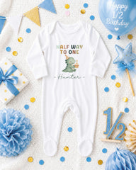 A white onesie with a dinosaur graphic and the text "Half Way to One Hunter" is displayed among various baby-related items, including a blue balloon, confetti, and a star-shaped decoration.