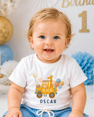 A smiling baby boy wearing a white t-shirt with a cartoon car and balloons, sitting in front of a birthday party backdrop.