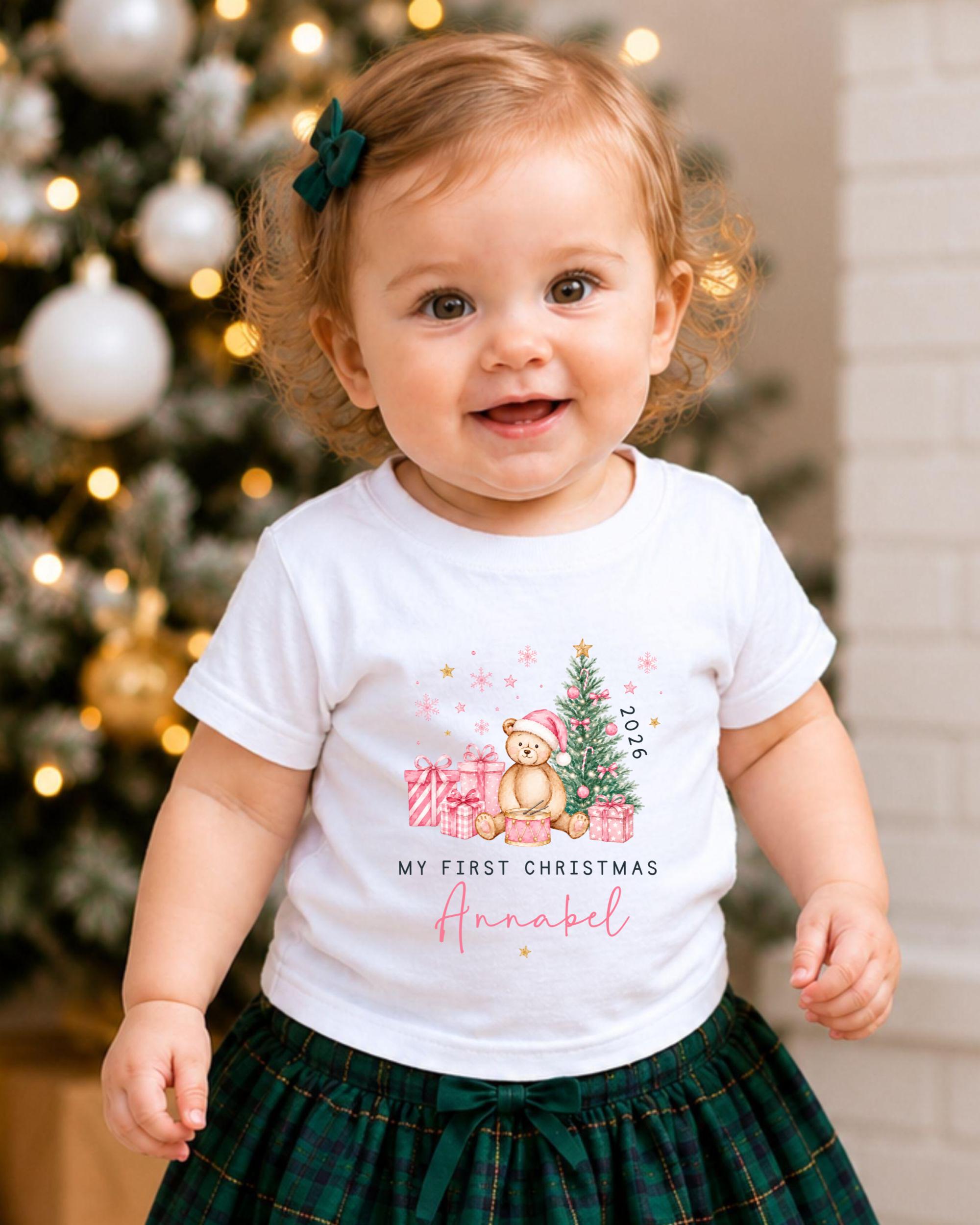 A smiling baby girl wearing a white t-shirt with a Christmas-themed design and a green bow in her hair, standing in front of a Christmas tree.