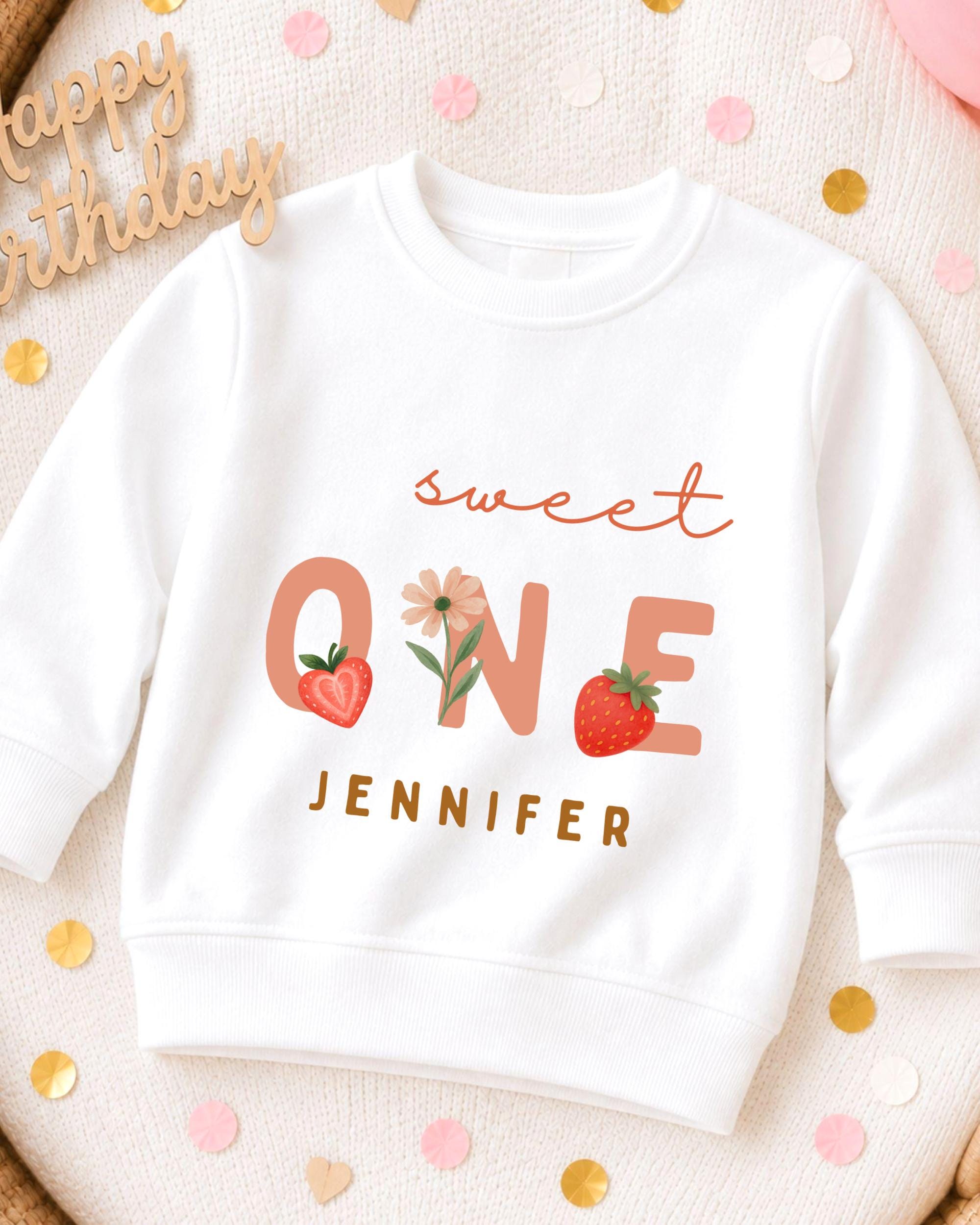 A white sweatshirt with a personalized text and graphics design, including the text "Sweet One" and a strawberry, along with the name "JENNIFER".