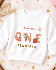 A white sweatshirt with a personalized text and graphics design, including the text "Sweet One" and a strawberry, along with the name "JENNIFER".