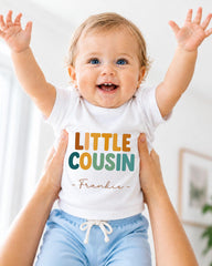 A smiling baby wearing a white shirt with the text "LITTLE COUSIN - Frankie" is being held up by someone's arms.