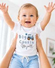 A smiling baby wearing a white shirt with a tractor graphic and the text "Big Cousin Matt" is being held up by someone's arms.