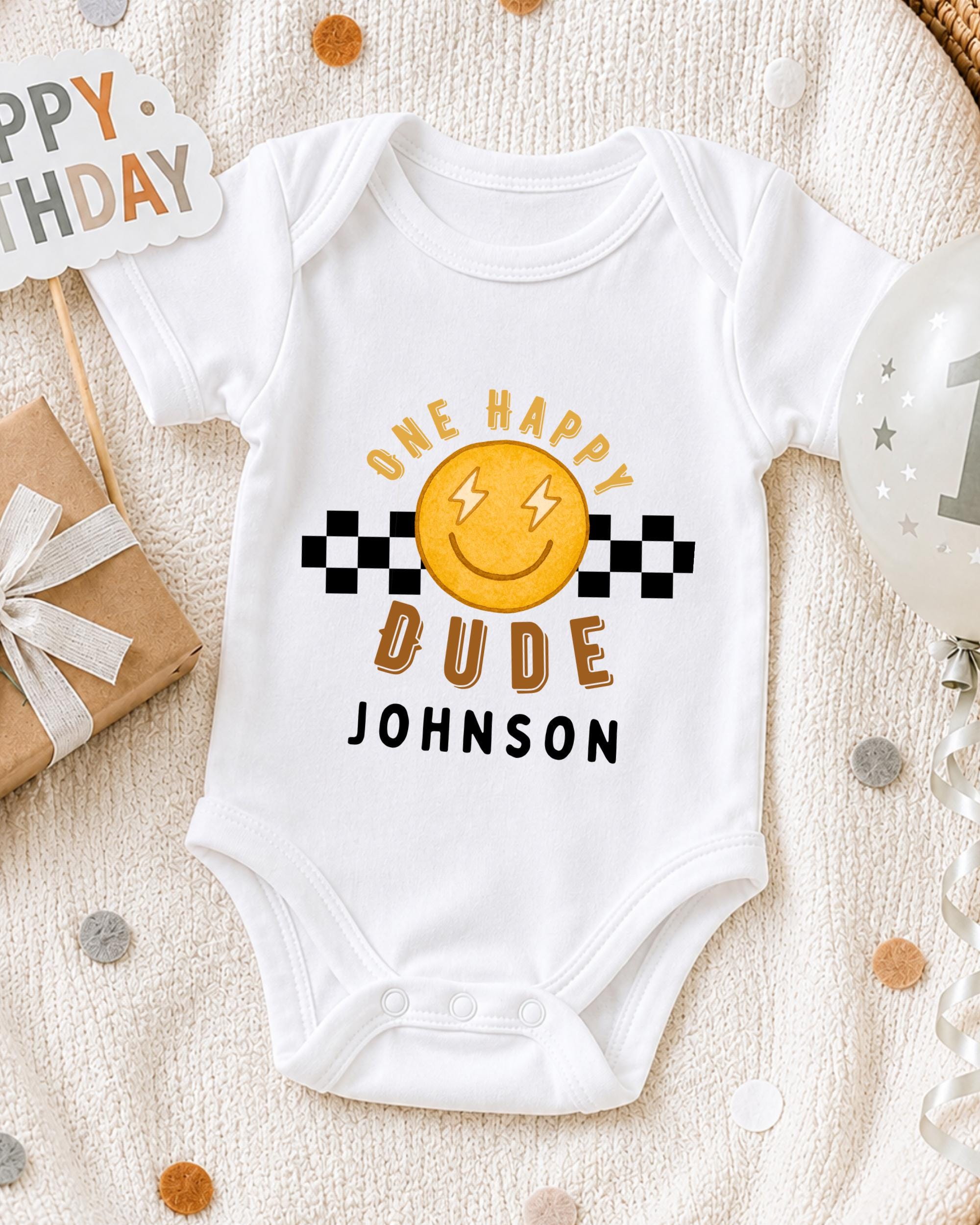 A white onesie with a smiley face and the text "ONE HAPPY DUDE JOHNSON" printed on it, along with a "Happy Birthday" sign and a birthday balloon.