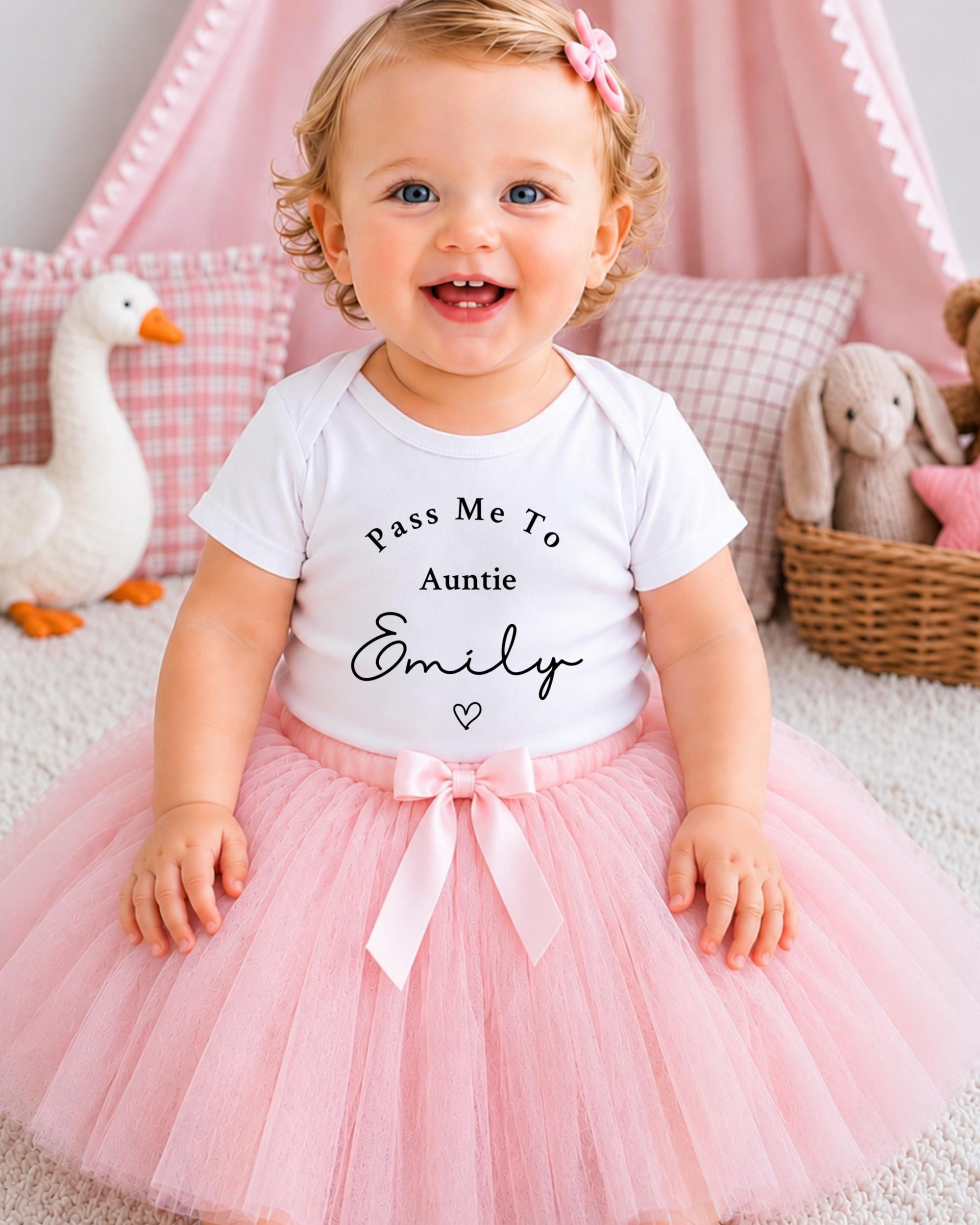 A smiling baby girl wearing a white t-shirt with the text "Pass Me To Auntie Emily" and a pink tutu skirt, sitting on a pink and white checkered rug.