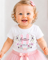 A smiling baby girl wearing a white t-shirt with a floral design and a pink bow in her hair.