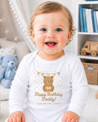 A smiling baby wearing a white onesie with a teddy bear and birthday decorations, sitting in a room with bookshelves in the background.