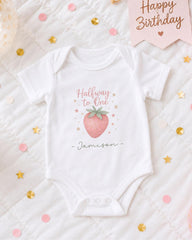 A white onesie with a pink strawberry and stars printed on it, placed on a bed with confetti and a birthday card.