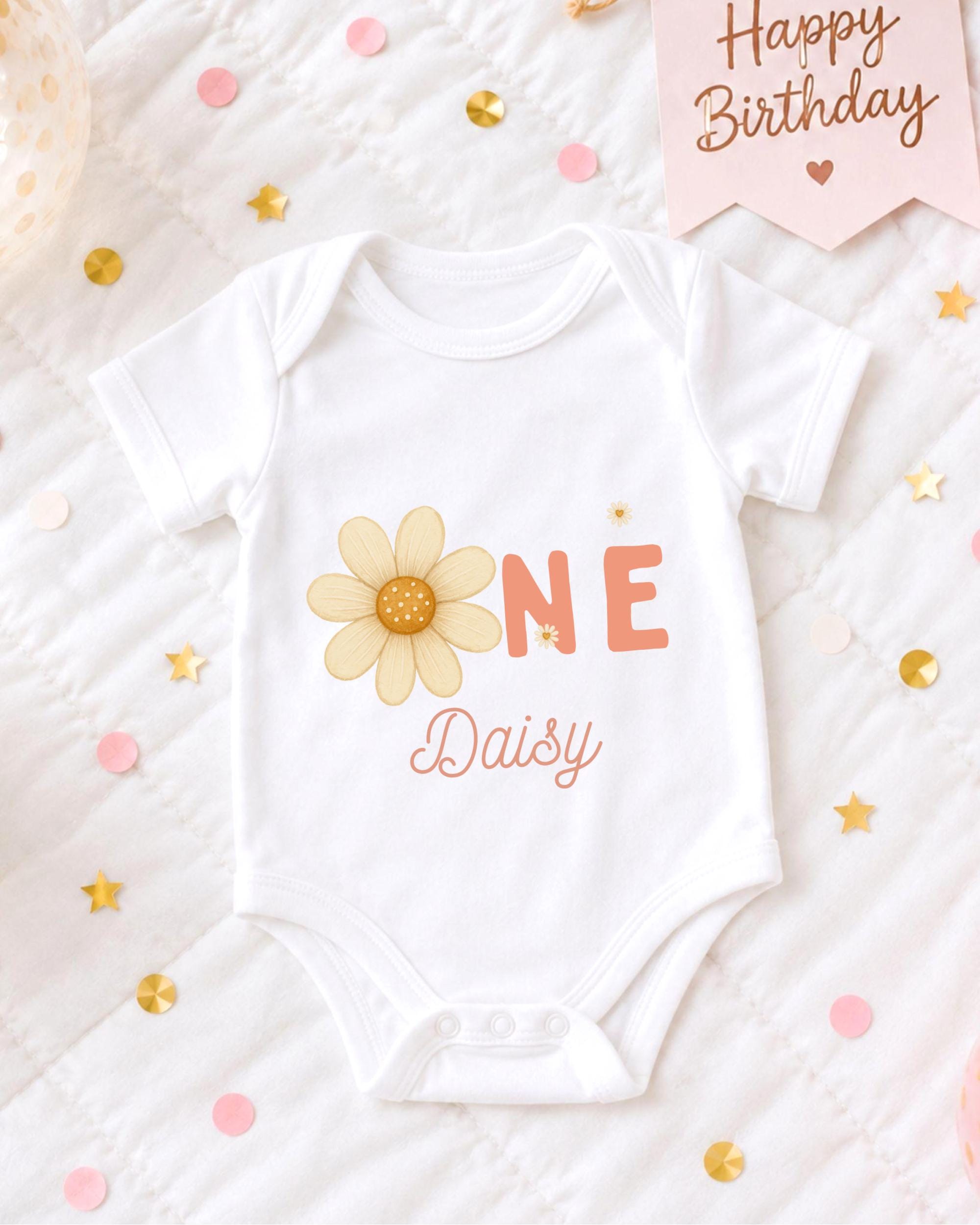 A white onesie with a flower design and the text "Daisy" printed on it, surrounded by confetti and a birthday card.
