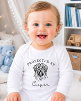 A smiling baby wearing a white onesie with a dog graphic and the text "PROTECTED BY Cooper" printed on it.
