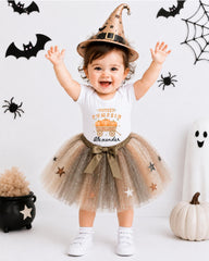 A young girl wearing a witch's hat and a tutu with stars on it, smiling and standing in front of Halloween decorations.