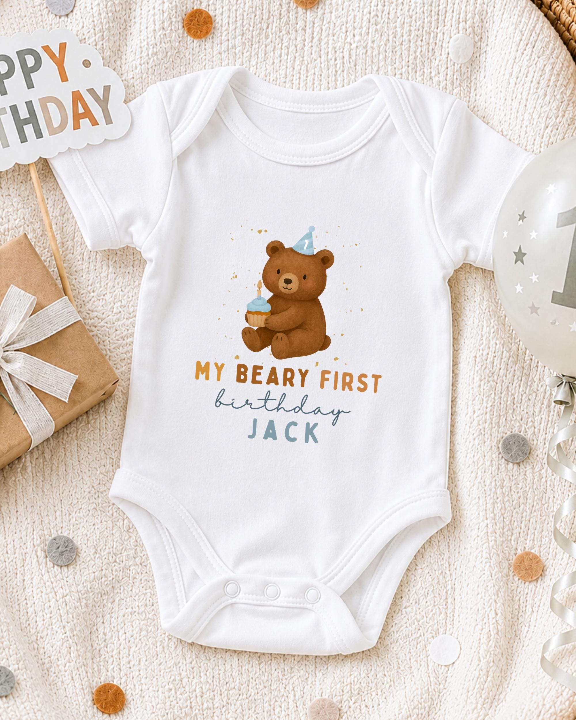 A white onesie with a bear and birthday text, along with a "Happy Birthday Jack" sign, a gift box, and a balloon, all arranged on a white blanket with confetti.