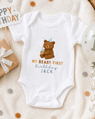 A white onesie with a bear and birthday text, along with a "Happy Birthday Jack" sign, a gift box, and a balloon, all arranged on a white blanket with confetti.