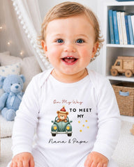 A smiling baby wearing a white onesie with a cartoon image of a bear driving a car and text that says "On My Way to Meet My Nana & Papa".