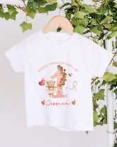 A white t-shirt with a personalized design hanging on a wooden hanger. The design features a teddy bear, a pink number one, and the text "When I wake up I will be Jessica".