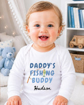 A smiling baby boy wearing a white onesie with the text "DADDY'S FISHING BUDDY" and the name "Hudson" printed on it.