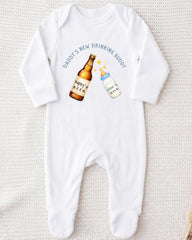 A white onesie with a graphic design on it, depicting a beer bottle and a baby bottle. The text above the graphic says "DADMY'S NEW DRINKING BUDDY".