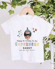 A white t-shirt with a printed design featuring a superhero character and text that reads "Happy Birthday to my SUPERHERO DADDY love you lots, Olilee".