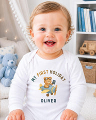 A smiling baby boy wearing a white onesie with a teddy bear and airplane graphic, sitting in a room with bookshelves in the background.