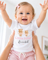 A smiling baby girl wearing a white t-shirt with a design of two teddy bears and the text "Little Sister" and "Annabel" printed on it.