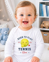 A smiling baby boy wearing a white onesie with a tennis design and text.