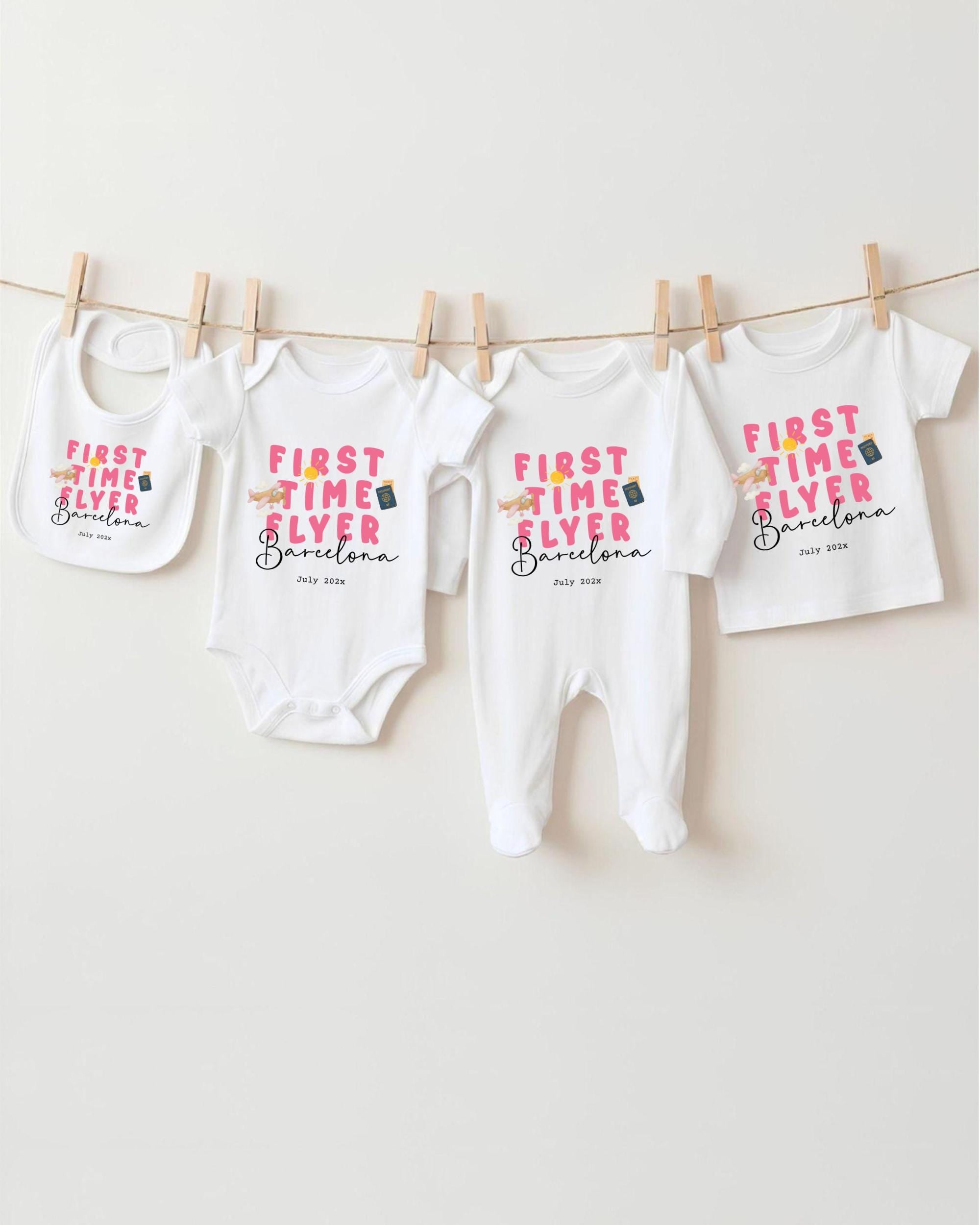 A row of four white baby clothes with the text "FIRST TIME FLYER Barcelona" printed on them, hanging on a clothesline.
