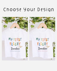 two white t-shirts with the text "My first flight Barcelona Custom Month, year" printed on them. The t-shirts are hanging on a wooden hanger, with a plant visible in the background.