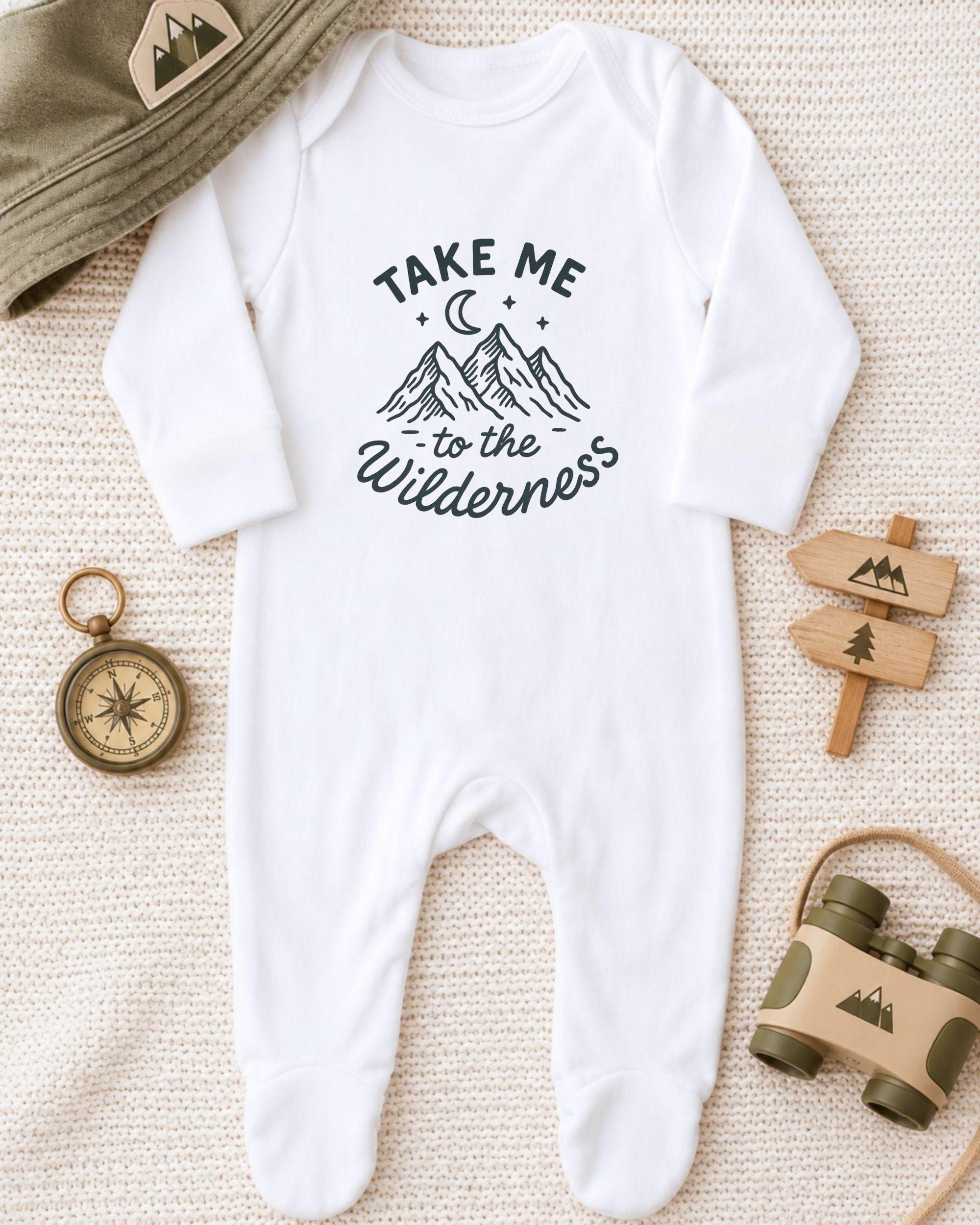A white onesie with a mountain and star design, along with a compass, a hat, and a toy binoculars, all arranged on a beige surface.