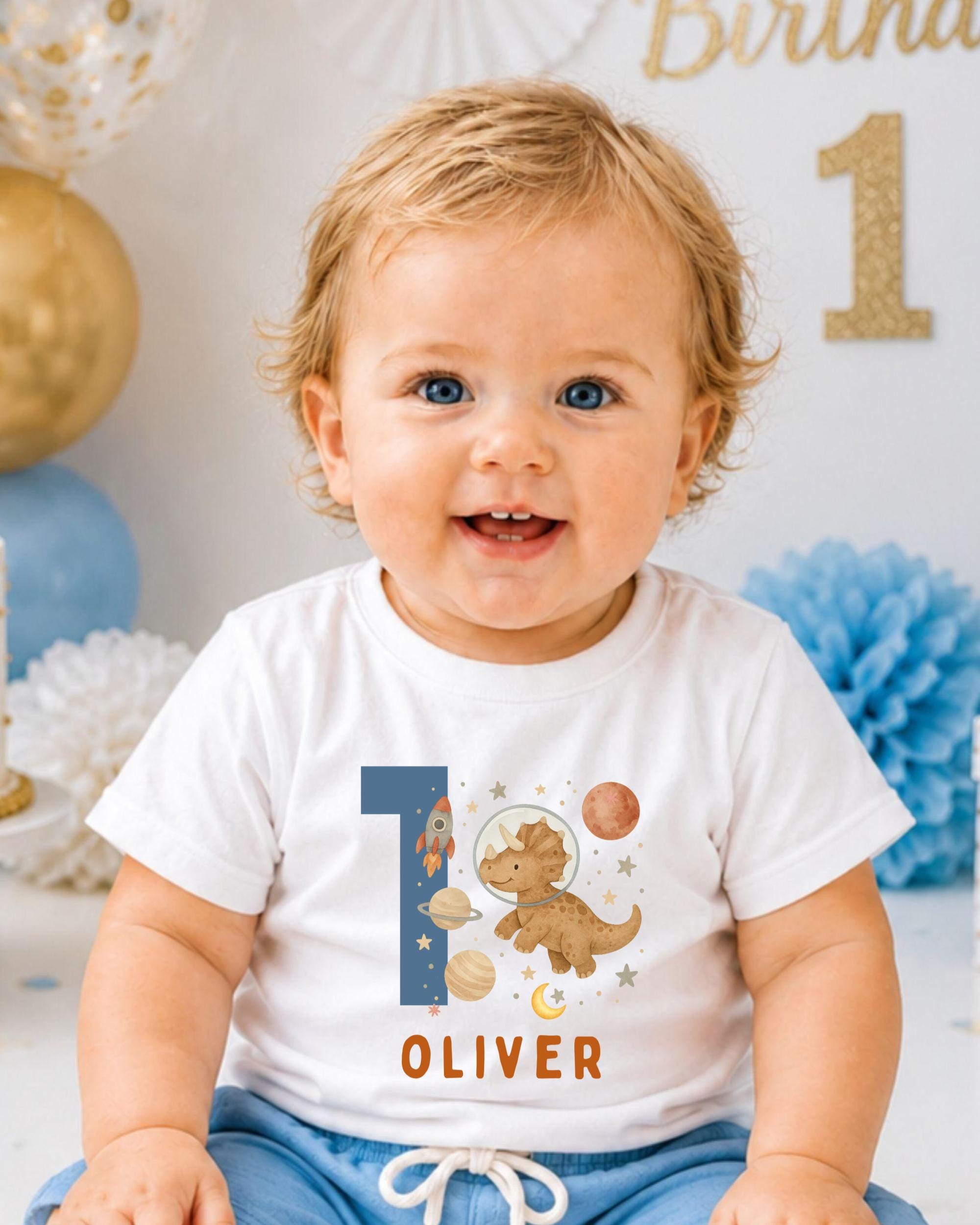 A smiling baby boy wearing a white t-shirt with a dinosaur and space-themed design, sitting in front of a birthday party backdrop.