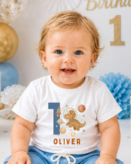 A smiling baby boy wearing a white t-shirt with a dinosaur and space-themed design, sitting in front of a birthday party backdrop.