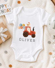 A white onesie with a red tractor and a chicken on a birthday-themed background, along with a "Happy Birthday" sign and a birthday balloon.