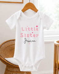 A white baby onesie with the text "Little Sister Jessica" printed on it, hanging on a wooden hanger.