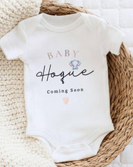 A white baby onesie with the text "BABY Hogue" printed on it, along with a small elephant image. The text "Coming Soon" is also printed below the image.