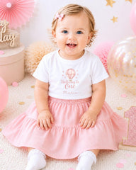 A smiling baby girl wearing a white t-shirt with a balloon design and a pink skirt, sitting on a pink and white background with balloons and confetti.