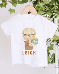 A white t-shirt with a printed design of a hot air balloon carrying a teddy bear and other animals, along with the name "LEIGH" printed below.
