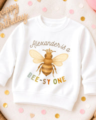 A white sweatshirt with a bee graphic and text printed on it, placed on a surface with confetti.