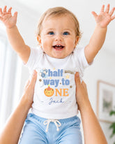A smiling baby wearing a white onesie with the text "Half Way to One Jack" is being held up by someone's arms.