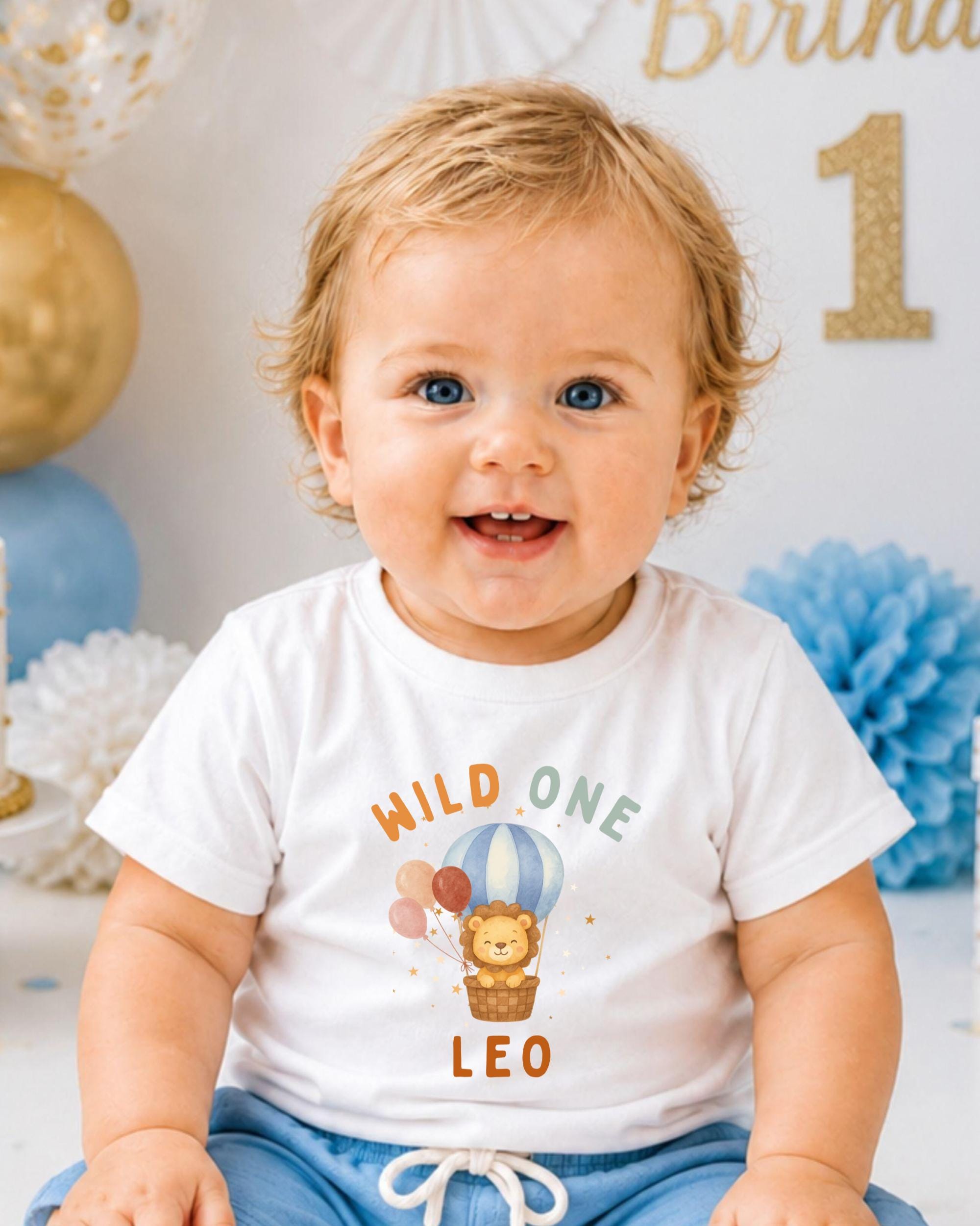 A smiling baby boy wearing a white t-shirt with a cartoon lion and a hot air balloon graphic, sitting in front of a birthday party backdrop.