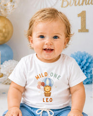 A smiling baby boy wearing a white t-shirt with a cartoon lion and a hot air balloon graphic, sitting in front of a birthday party backdrop.