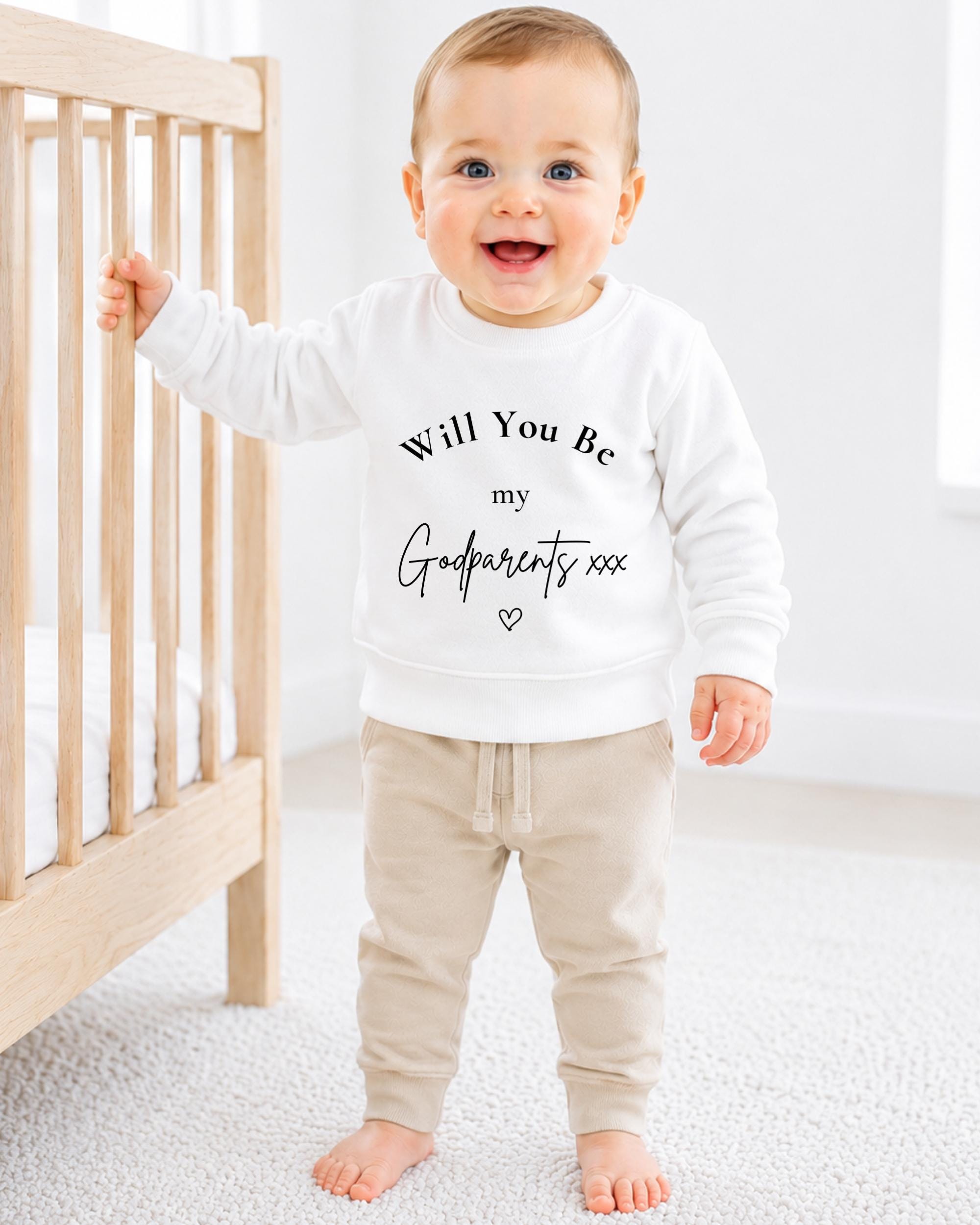 A smiling baby wearing a white sweatshirt with the text "Will You Be my Godparents xxx" printed on it.