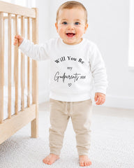 A smiling baby wearing a white sweatshirt with the text "Will You Be my Godparents xxx" printed on it.