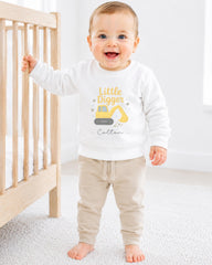 A smiling baby boy wearing a white sweatshirt with a yellow construction vehicle graphic and the text "Little Digger" is standing next to a wooden crib.