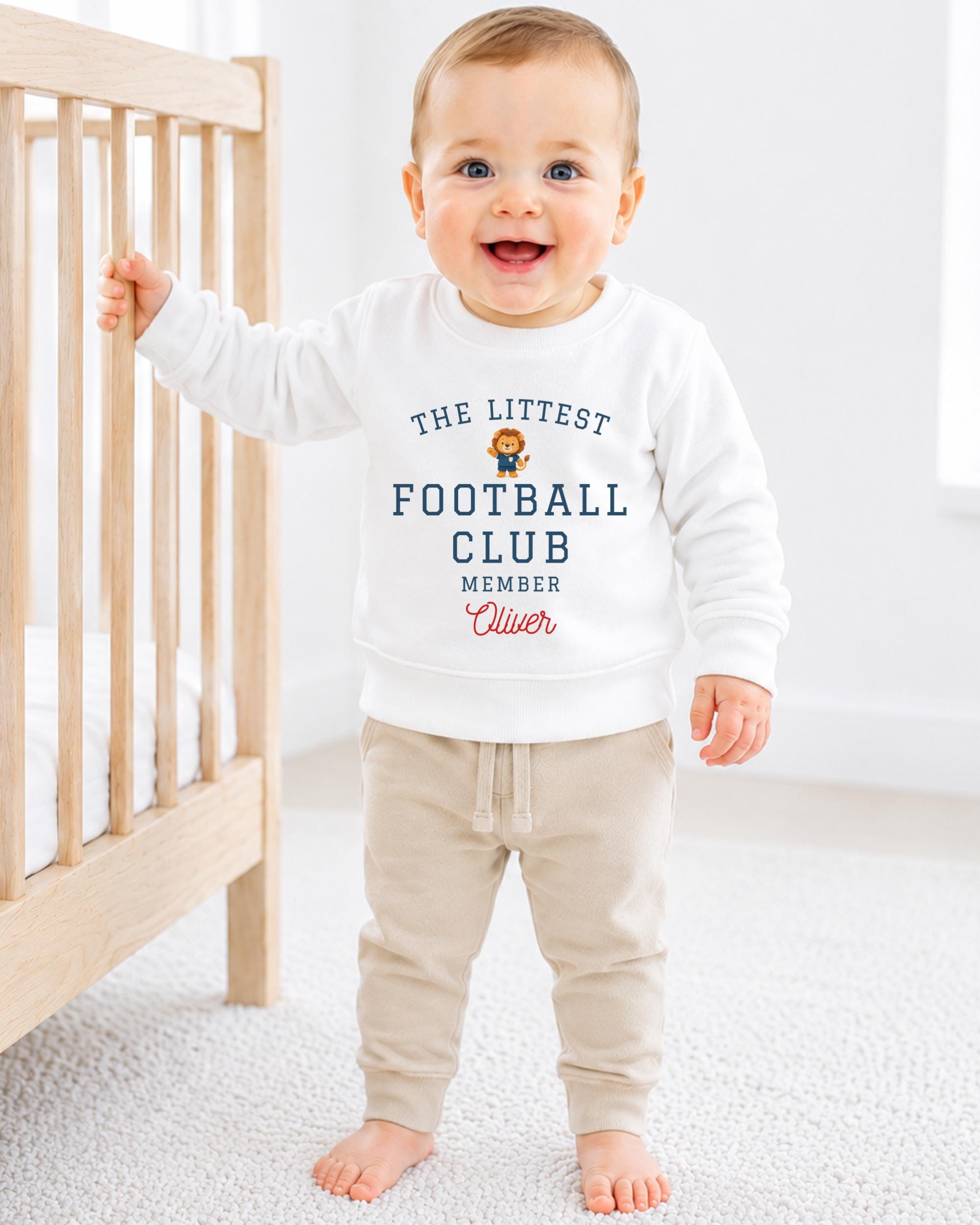 A smiling baby boy wearing a white sweatshirt with text and a logo, standing in front of a wooden crib.